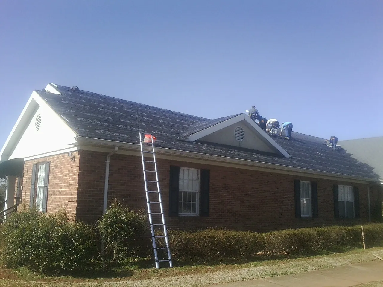 Crew tearing shingles off a brick ranch roof with a ladder set — Metal Roof Repair work by our crew in Pewamo