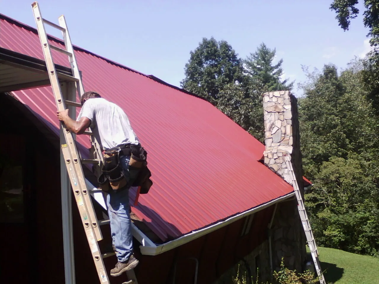 Roofer on a ladder beside a red metal roof and stone chimney — Soffit & Fascia Repair by Roofing Brothers in Pewamo